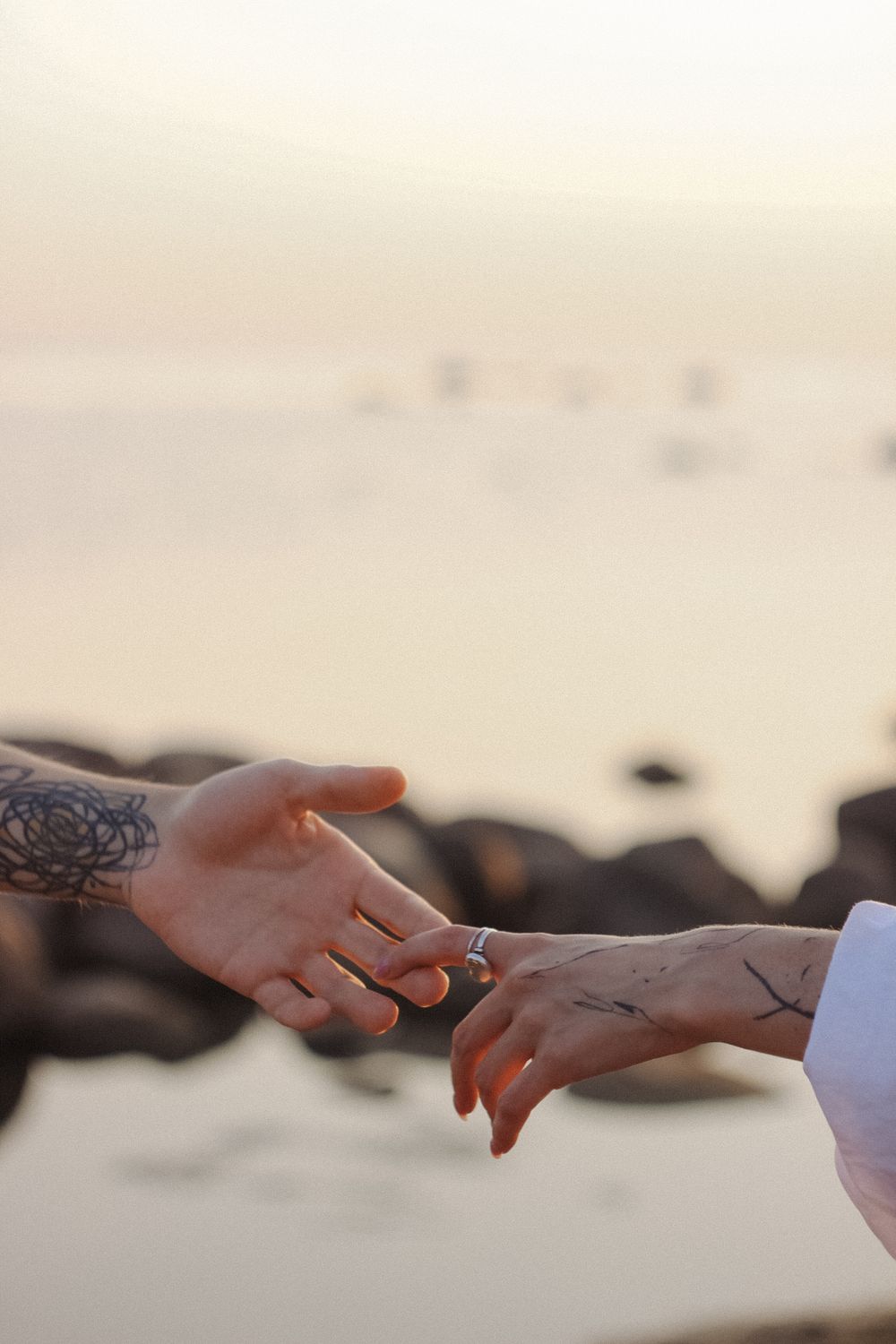 Close-up of a couple's hands reaching for each other by the sea at sunset, symbolizing love and connection.