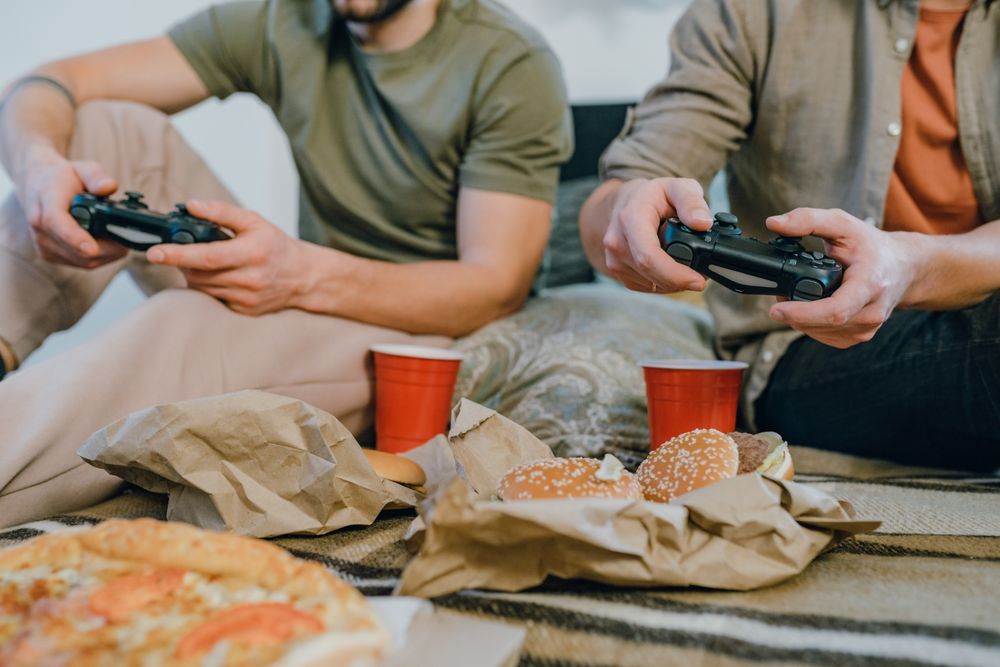 Two friends playing video games and enjoying burgers and pizza indoors with red cups.